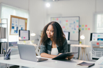 Young african american manager is sitting at her desk and working on a laptop while holding a report in her other hand. She is smiling and looking at the laptop screen