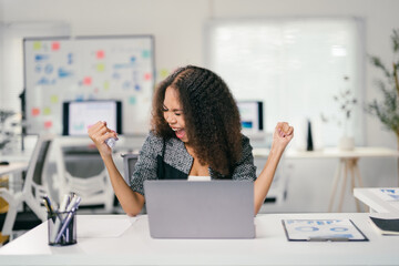 Enthusiastic young office worker is raising her fists in the air and cheering while sitting at her...