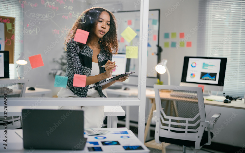 Poster young businesswoman is analyzing data and working on new project, using sticky notes on a glass boar