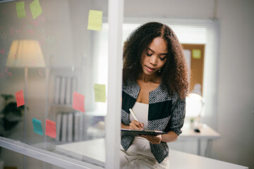 Young businesswoman is standing in an office setting, taking notes on a clipboard while reviewing information on sticky notes stuck to a glass partition