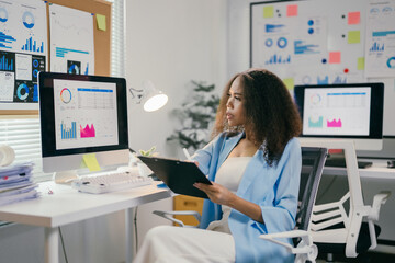 Focused businesswoman analyzing financial data in office, taking notes on clipboard surrounded by charts and graphs, showcasing expertise in data analysis and decision-making