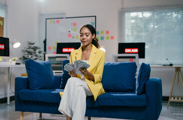 Young businesswoman sits on a blue sofa, counting money in a modern office, exuding confidence and...