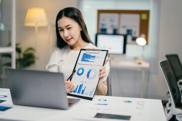 Businesswoman is presenting charts and graphs on a video call from her brightly lit office at night. She is pointing to a chart with a pen while looking at her laptop