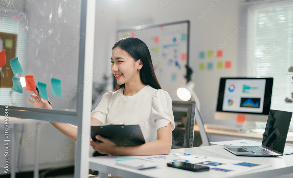 Aufkleber young manager is smiling while planning a project using adhesive notes on a glass wall in her office