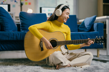 Young woman is sitting on the floor in her living room, playing an acoustic guitar and wearing headphones. She is smiling and enjoying her music