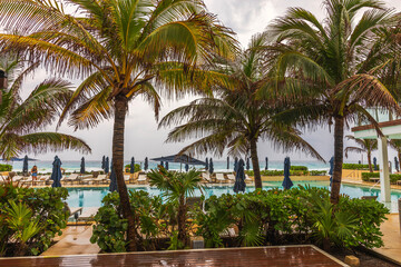 Fototapeta premium Poolside view with palm trees and lounge chairs in tropical resort by Caribbean sea under overcast skies. Mexico.