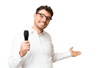 Brazilian man picking up a microphone over isolated chroma key background extending hands to the side for inviting to come