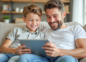 father and son are sitting on the couch looking at a tablet and laughing