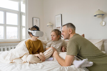 A father and his two daughters are spending time together on the bed, the older daughter is wearing a VR headset