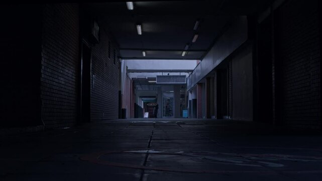 Moody footage of a dark, covered alley in a semi-derelict UK shopping centre. Dull fluorescent lights and cold autumnal weather create a tense atmosphere suiting a crime theme.