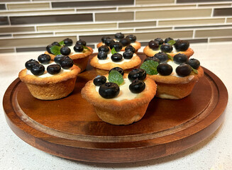 A batch of freshly made blueberry tartlets sit beautifully on a wooden tray. Each tartlet has a golden-brown crust, filled with smooth vanilla custard, and is topped with plump blueberries.