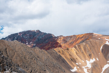 Colorful layered landscape with sunlit sharp rocky mountain ridge of vivid colors under cloudy sky. Golden rocks and red sheer crags in sunlight. Shadows of clouds on unusual multicolored mountains.