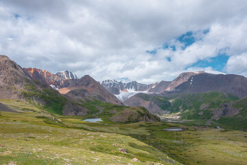 Colorful scenery with two beautiful lakes among green hills and rocks in alpine valley with view to big glacier and large snow mountain range far away under clouds in blue sky. High snowy mountains.