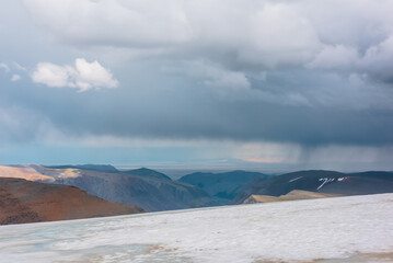 Dramatic alpine top view from flat sloping glacier on precipice edge to deep gorge between colorful sunlit cliffs and mountain range silhouette in rain under lead gray cloudy sky. Rainy grey clouds.