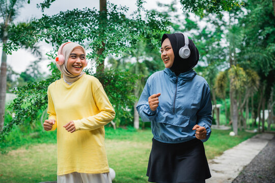 Two women wearing headphones are happily running outdoors in a lush green park setting - Powered by Adobe