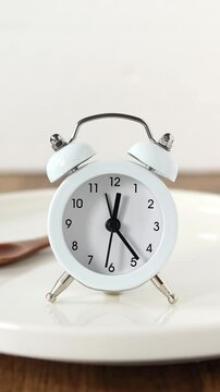 Alarm clock on empty plate and wooden spoon with white background. Concept of intermittent fasting and meal planning.