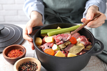 Woman preparing stew with vegetables and meat at white tiled table, closeup
