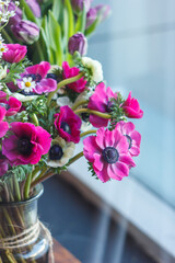 A vibrant bouquet of pink anemones in a glass vase, captured in natural light by a window, showcasing fresh, vivid blossoms and soft greenery in an elegant floral arrangement.