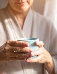 Mature caucasian woman is drinking tea or coffee from cup near window at home. Concept of domestic lifestyle. Cropped photo of modern happy middle aged lady wearing bathrobe at sunny day. 