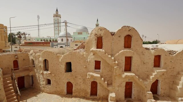 KSAR HADADA, TUNISIA - MARCH 03, 2024: Scenic view of ancient weathered Berber earthen ghorfas contrasting with modern mosque in background under warm Tunisian spring sun in village of Ksar Hadada
