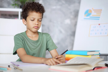 Portrait of adorable African American schoolboy sitting at the table with his homework, looking unhappy andtired unwilling to continue studying