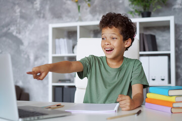 Excited African American kid student pointing with his index finger at the laptop computer screen with "wow" open mouth face expression during his online lesson