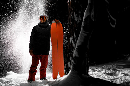 Young male snowboarder in ski goggles holds his red splitboard next to him, standing near trees