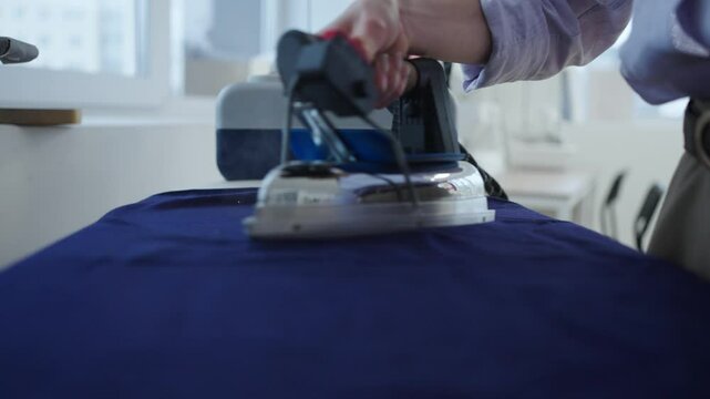 Close-up view of a seamstress ironing blue fabric with a professional iron in a sewing workshop, showcasing the meticulous process of preparing textiles for garment creation