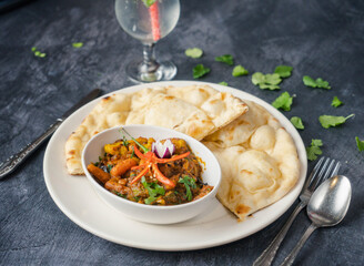 Special meat curry in a bowl surrounded by fluffy naan bread. The dish is garnished with fresh herbs and colorful chili slices, and a refreshing drink with mint and lime is beside the plate.