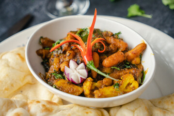 A bowl of vibrant curry sits at the center, adorned with colorful spices and garnished with fresh herbs and a sliced red chili. Surrounding the dish are warm, fluffy naan bread.