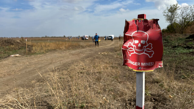 danger sign mine in Ukrainian near an agricultural field during war in Ukraine, with people from demining team in background. Translation: "Mine Danger"