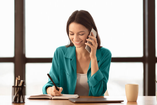 Smiling business woman talking on phone and taking notes, sitting at her working place in office, copyspace - Powered by Adobe