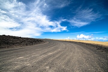 A dirt road curves gently across a rugged volcanic terrain, with vibrant blue skies and wispy clouds overhead