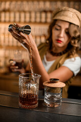 Focus on a mixing glass on the bar, into which a female bartender pours a brown drink from a jigger