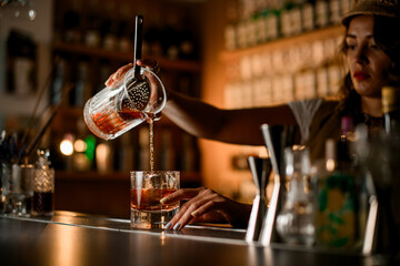 Female hands pouring a cocktail from a mixing glass into a glass with an ice cube