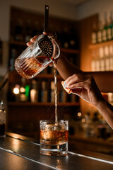 On the bar stands a glass with an ice cube, into which female hands pour a brown cocktail from a...