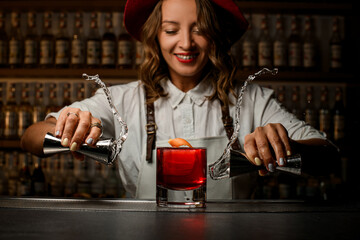 Happy female bartender holds a jigger with a transparent drink in both hands, which she pours on each other