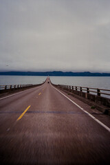 Narrow Bridge Surrounded by Water Leading to Distant Green Landscape Under a Stormy Sky