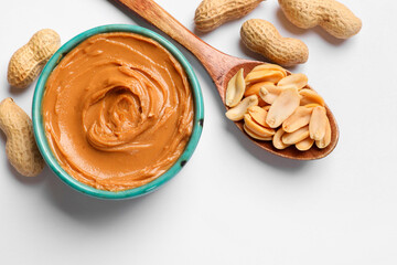 Tasty peanut butter in bowl and spoon with groundnuts on white table, flat lay