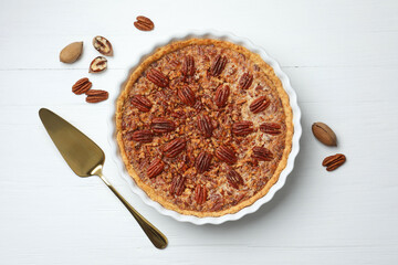 Delicious pecan pie in baking dish, cake server and fresh nuts on white wooden table, flat lay