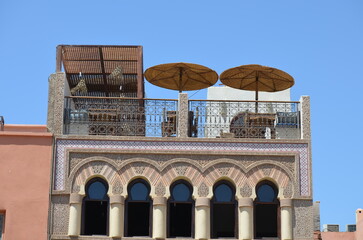 Obraz premium Traditional Moroccan terrace with decorative arches, ornate ironwork, and woven parasols in Marrakech, under clear blue sky 