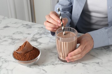 Woman with glass of protein cocktail and powder at white marble table, closeup
