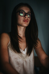 A young woman with long hair and glasses looks intently at a computer screen, reflecting...