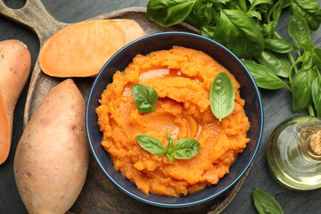 Tasty mashed sweet potato with basil in bowl, fresh vegetables and oil on dark gray table, flat lay