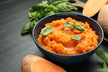 Tasty mashed sweet potato with basil in bowl and fresh vegetables on dark gray textured table, closeup
