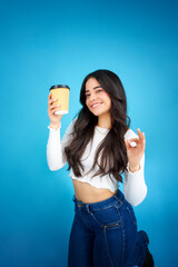 Happy young latin woman showing disposable coffee cup doing ok sign with hand in studio