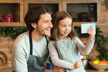 Weekend With Daddy. Cheerful Father And Little Daughter Taking Selfie On Smartphone In Kitchen, Having Fun While Cooking At Home, Preparing Healthy Food, Wearing Aprons, Smiling At Camera, Free Space