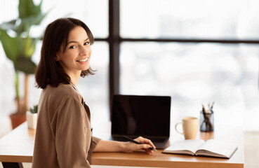 Online Education. Smiling young teacher holding pen looking at camera, sitting at desk. Laptop mock up, empty space