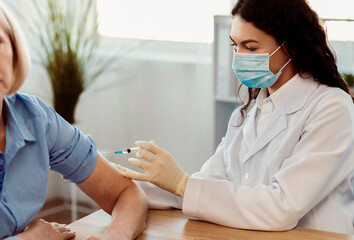 Seasonal Flu Shot. Mexican doctor in medical mask injecting tetanus toxoid vaccine to mature woman,...