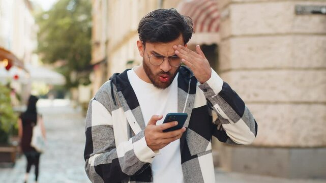 Indian young man using smartphone typing browsing, loses becoming surprised sudden lottery results, bad news, fortune loss, fail. Guy tourist standing on urban city street outdoors. Town lifestyles.